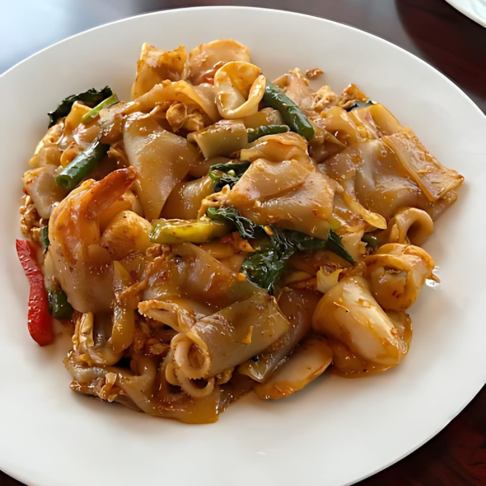 Close-up of a Thai dish with wide noodles, shrimp, squid, and green vegetables on a white plate.