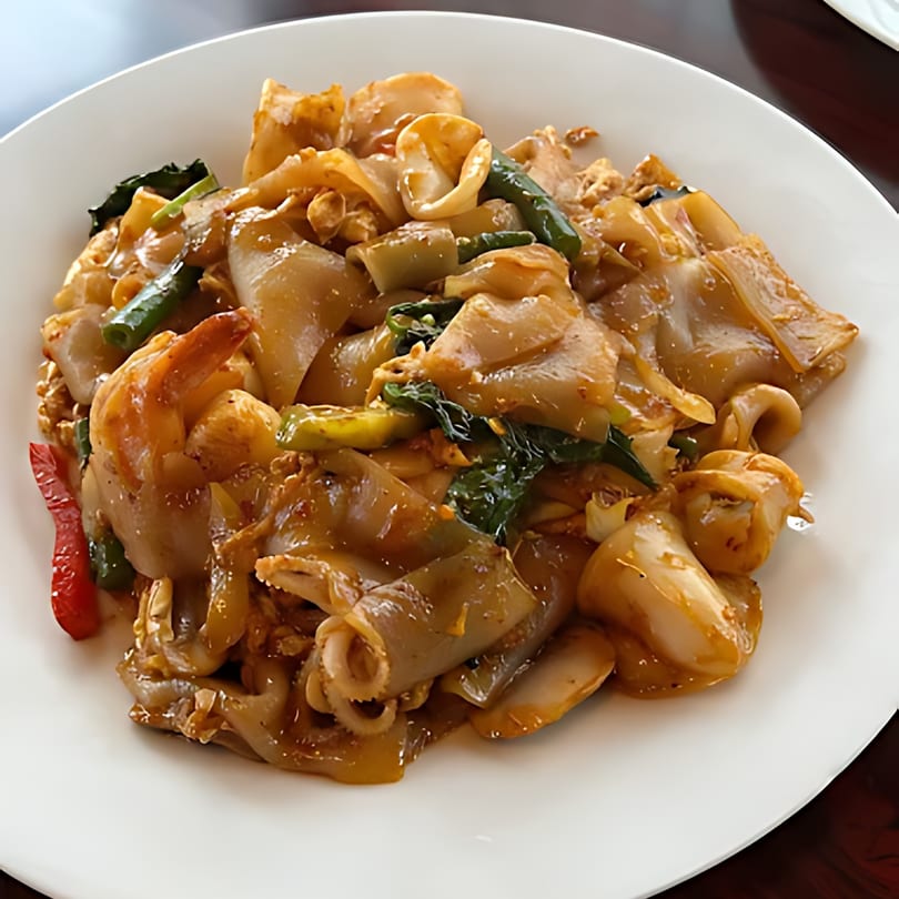 Close-up of a Thai dish with wide noodles, shrimp, squid, and green vegetables on a white plate.