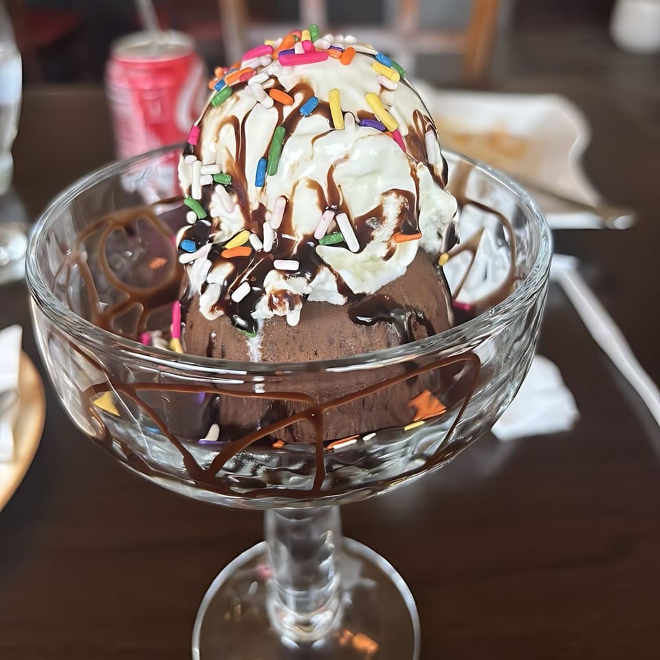 Chocolate ice cream sundae with whipped cream, chocolate syrup, and colorful sprinkles in a glass dish.